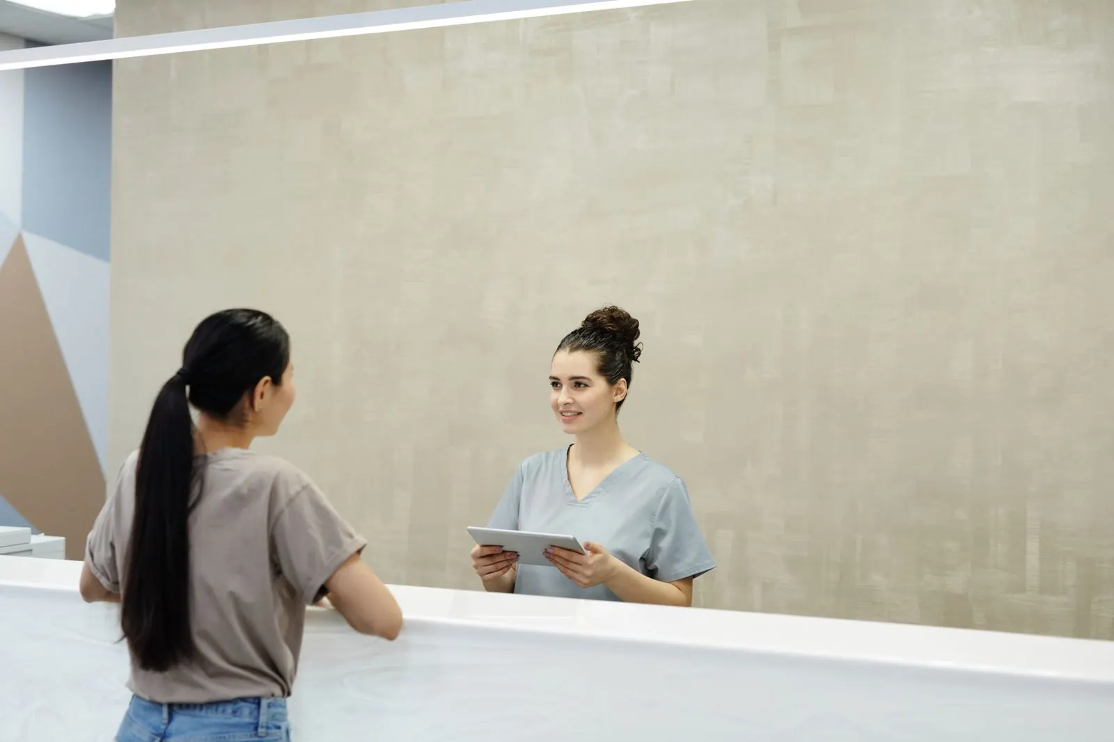 A receptionist with a tablet greeting a visitor at a clinic front desk